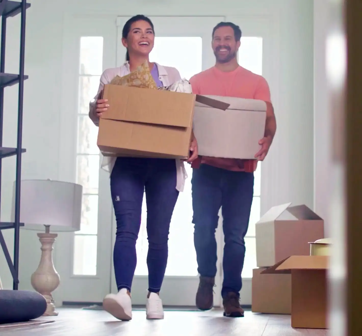 Two adults have just entered their new home holding boxes and smiling.