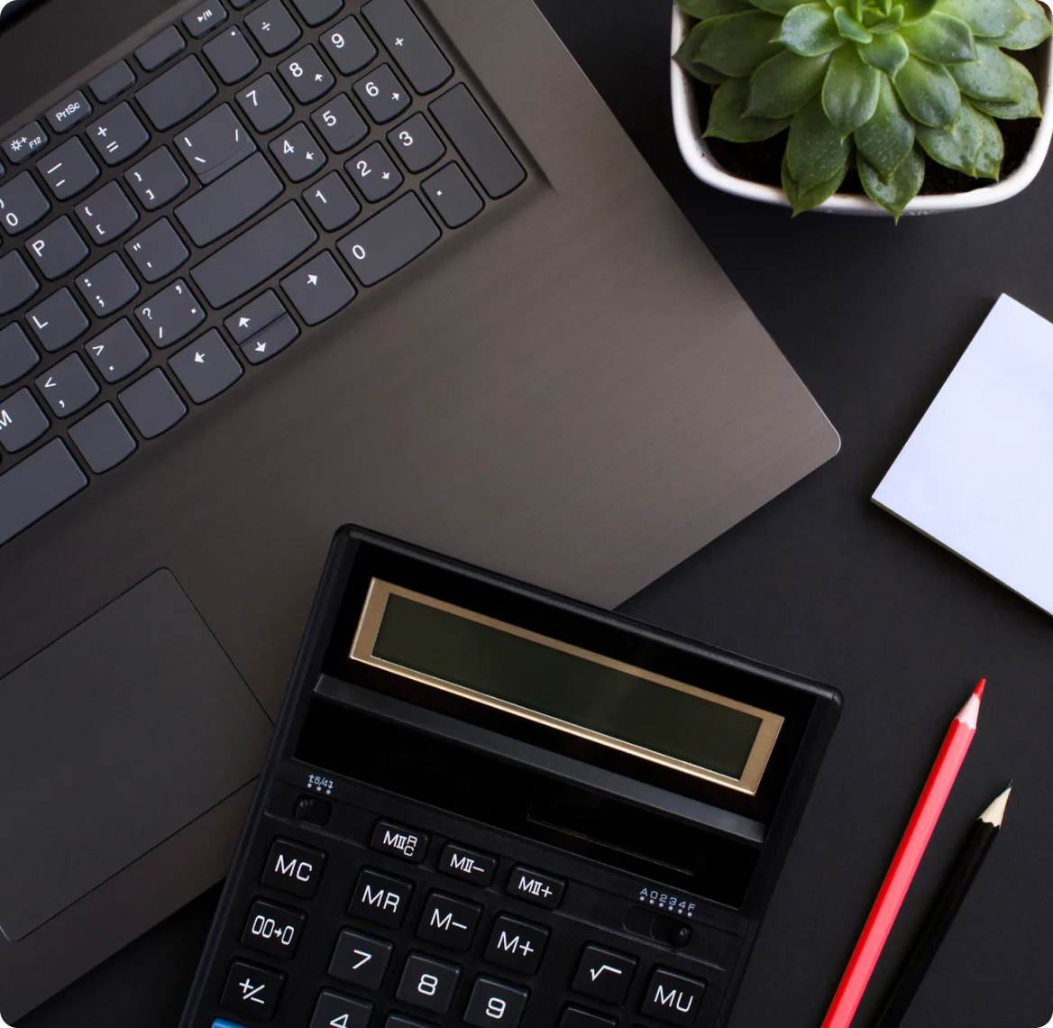 A desktop with a laptop, calculator, red and black colored pencils, a sticky note and a small potted plant.