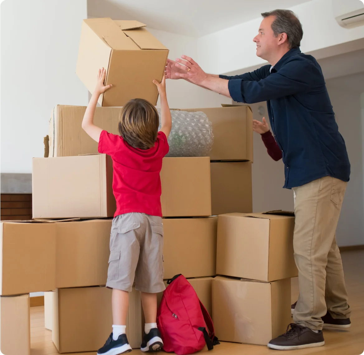 A father in tan pants and blue long-sleeved shirt helps his young son in a red shirt and gray shorts place a box on top of a stack of unmarked boxes.