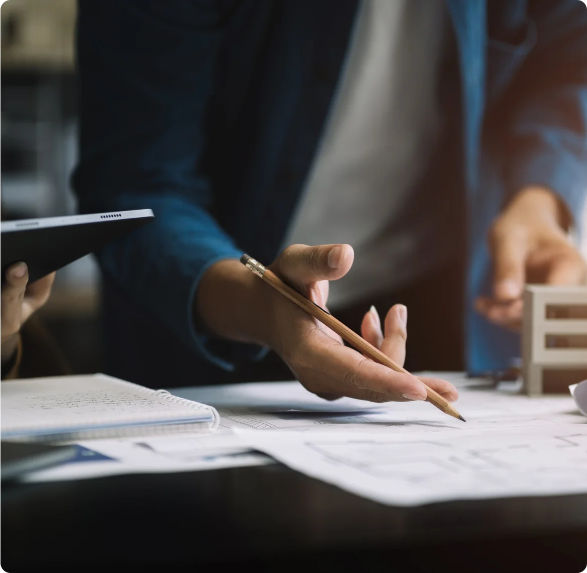 2 people mid-discussion reviewing planning documents. The image is cropped such that the focus is on one of the individual's hands, holding a pencil, hovering over the documents.