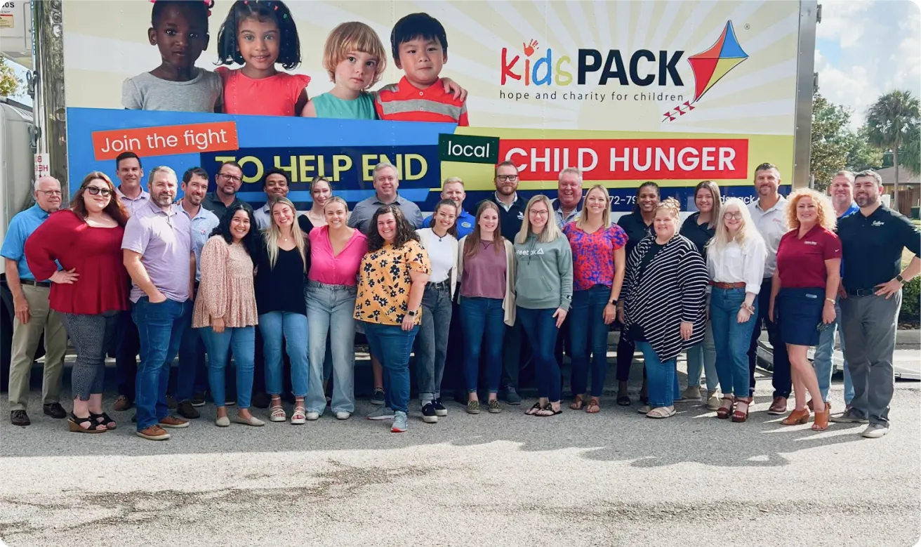 A group of people standing in front of a sign that reads 'KidsPACK home and charity for children. Join the fight to help end local child hunger.'