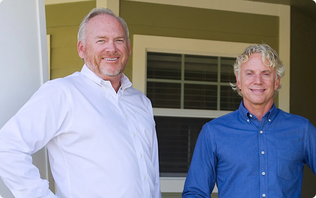 2 smmiling men standing in front of a home. The man on the left is taller, has beard stubble with light gray hair and is wearing a white collared button-down shirt. The man on the right is clean-shaven, shorter with curly blond hair and wearing a blue collared button-down shirt.